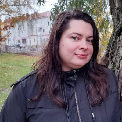 Nainen nojaa puuhun pihamaalla ja katsoo kameraan.