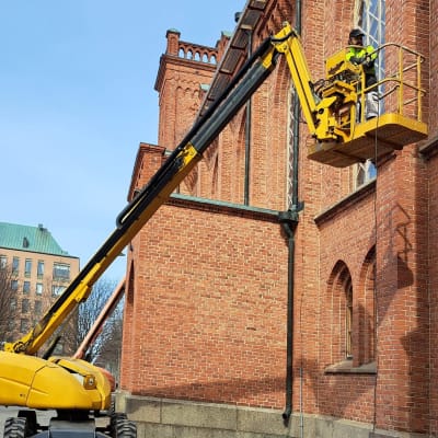 Rakennusmies nosturin nokassa korjaamassa Vaasan kirkon julkisivua. Mies valvoo maan tasalta.