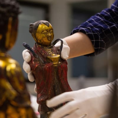 Aasialaisia taide-esineitä. Museon työntekijä nostaa yhtä pientä patsasta hansikkaat kädessä.