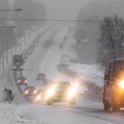 Bilar med lyktorna påslagna kör på en landsväg i snöväder.