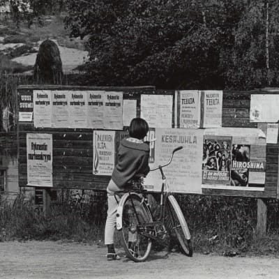 Maaseutu. Tyttö ja polkupyörä ilmoitustaulun edessä v. 1964. Elokuvamainoksia ja ilmoituksia. Kesä.