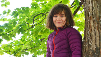 En ung flicka uppflugen i ett träd. Hon tittar in i kameran och ler.