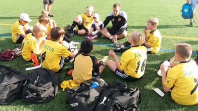 En grupp barn sitter i en ring tillsammans med tränare Markus Dahlbom på en fotbollsplan.