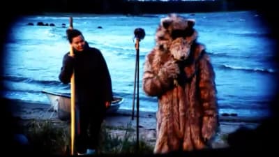 En fiskare och en person klädd i björnkostym står på en sandstrand. En liten roddbåt är uppdragen i strandvattnet.