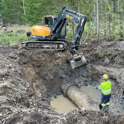 Kaksi työmiestä seisoo mutaisessa montussa, jossa on paksu putki.
