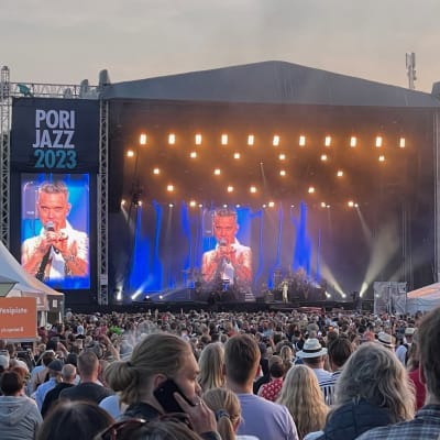 Valtava yleisö seisoo ison esiintymislavan edessä katsomassa Robbie Williamsin konserttia, Williamsin kuva on heijastettu lavan näytöille.
