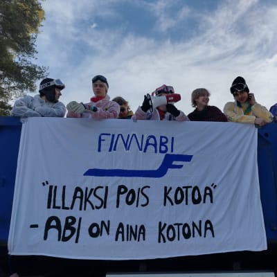 Ungdomar på lastbilsflak i Virkby, de är klädda i skiddress för utförsåkning . På affisch står det på finska (översatt) FinnAbi, hemifrån på kvällen, abirna är alltid hemma.
