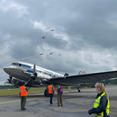 Vanha hopeinen DC-3 lentokone kiitoradalla. Taustalla taivaalla Sveitsin ilmavoimien taitolentohävittäjiä.