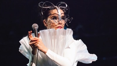 Artisten Björk med mikrofon i handen på scen i vita kläder och konstiga smycken i ansiktet.