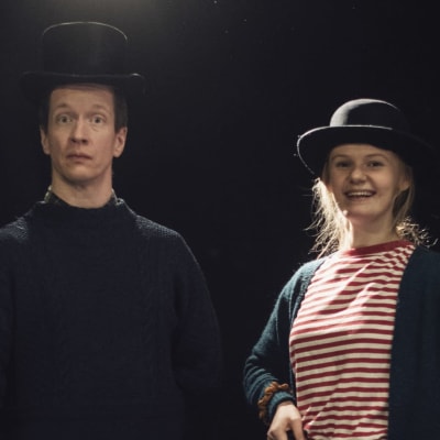Markus Haakana och Emma Nordblad står på en mörk scen med höga svarta hattar på huvudet.