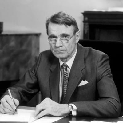 President Mauno Koivisto sitter vid ett bord och undertecknar ett papper.