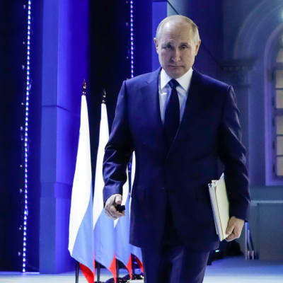 Vladimir Putin på väg bort från en talarstol. Han har ryska flaggor vid sin sida.