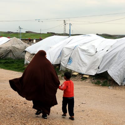 Hunnutettu nainen taluttaa pientä poikaa pakolaisleirin telttojen vieressä.