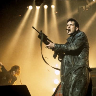 Mies mustassa nahkatakissa eli The KLF:n Bill Drummond lavalla konekivääri kädessä.
