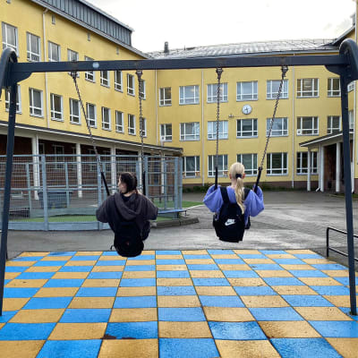 Kaksi yhdeksäsluokkalaista keinuu keltaisen koulun sisäpihalla.