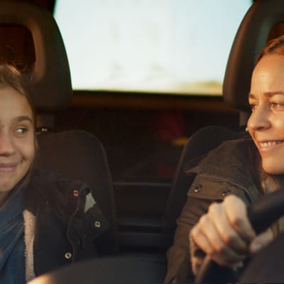 mor och dotter ler mot varandra i framsätet på en bil.