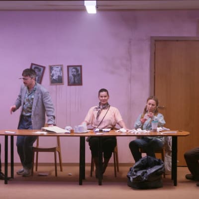 Fem skådespelare sitter bakom ett långt mötesbord i ett sjabbigt kontor. 