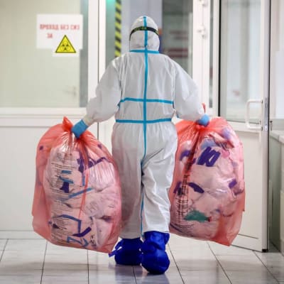 En person med skyddsutrustning bär två stora plastpåsar med avfall i händerna. Personen går mot en dörr i en sjukhuskorridor.