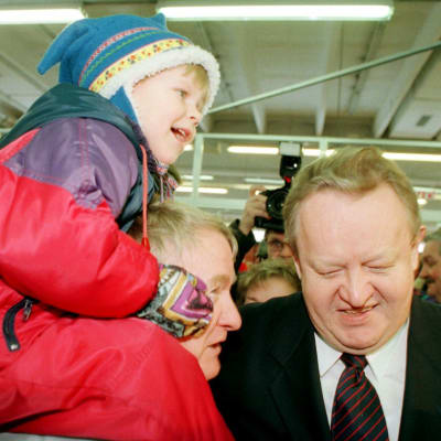 Presidentti Martti Ahtisaari katsoo alas nauraen, vieressä harmaapäinen mies, jonka harteilla poika punaisessa toppahaalarissa ja lapinlakissa, taustalla väentungosta.
