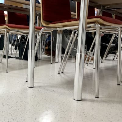 Luentosalin tuoleja ja pöytiä sekä niiden seassa opiskelijoiden jalkoja alhaalta kuvattuna.