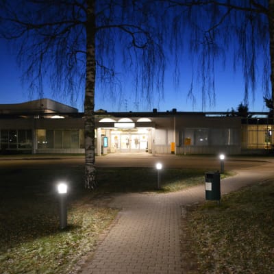 På bilden syns ingången till Borgå sjukhus om kvällen. Dörrarna finns på mitten av det avlånga huset. Den mörkblå kvällshimlen och några björkkvistar som hänger ner synns uppe på bilden.