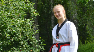 En ung kvinna i taekwondodräkt tittar in i kameran. Hon står utomhus framför ett äppelträd.