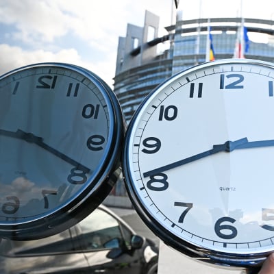 Käsi pitää kelloa ja Euroopan parlamenttirakennus Strasbourgissa siintää taustalla.
