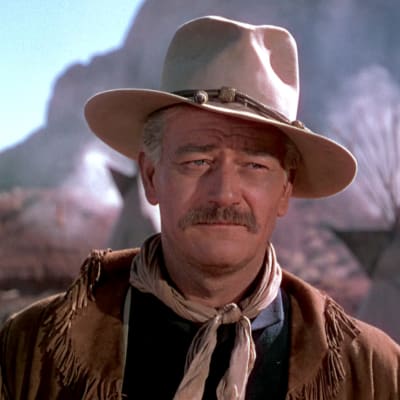 John Wayne hattu päässä, huivi kaulassa ja mokkanahkainen takki harteillaan. Taustalla näkyy intiaanikylä. Kuva elokuvasta Keltainen nauha.