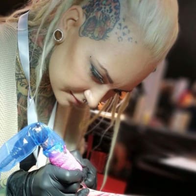 Tatuoija tekee asiakkaalleen tatuointia. Tatuoija on vaaleahiuksinen nainen, jolla on vaalea neuletakki ja tatutointeja rintakehässään, kaulassaan ja ohimollaan.
