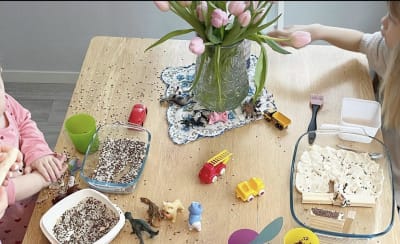 Två barn leker med så kallade "sensory bins", det vill säga upptäckarlådor. Bilden är tagen uppifrån på ett köksbord fullt med kärl fyllda av riskorn, träklossar och plastleksaker.