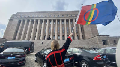En person i samiska kläder höjer en samisk flagga utanför riksdagshuset i Helsingfors.
