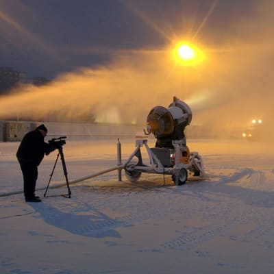 Kuvaaja kuvaa jalustakameralla urheilukentän reunassa lumitykin puhaltamaa lumipilveä aurinkoa vasten