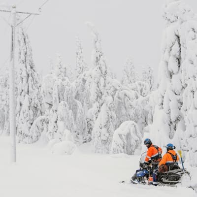 Två elreparatörer på snöskoter i en snöig skog.