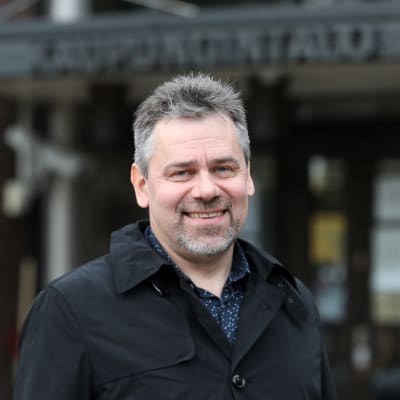 Mustaan takkiin pukeutunut Aki Viitasaari seisoo kaupungintalon edessä ja katsoo hymyillen kameraan.