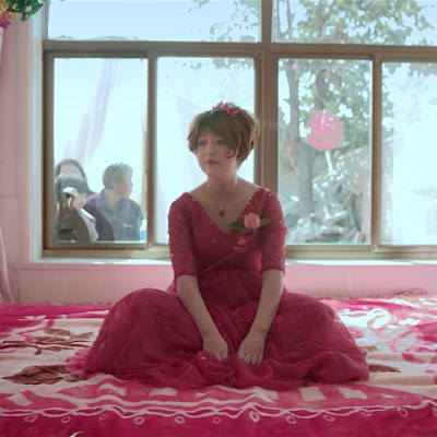 En kinesisk kvinna i röd bröllopsklänning sitter på en säng. I bakgrunden kikar nyfikna in genom fönstret.