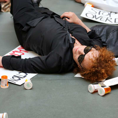 Närbild på Nan Goldin då hon ligger på golvet i ett museum, omgiven av en massa tomma pillerburkar och flygblad.
