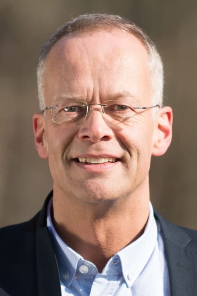 En man i blå skjorta och mörkare blå kavaj med glasögon utan skalmar. 