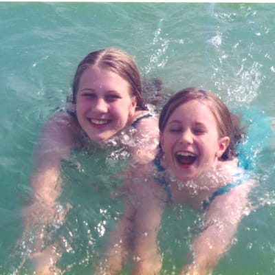 Kaksi tyttöä ui vihreässä vedessä ja nauraa kohti kameraa.