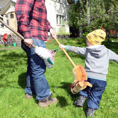 Isä ja lapsi leikkivät keppihevosilla ulkona.