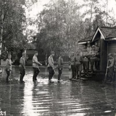 Mustavalkoisessa kuvassa varusmiehet seisovat jonossa isossa lätäkössä.