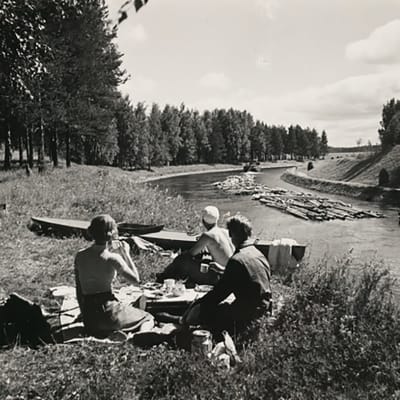 Svartvit bild: Tre personer har piknik vid kanalstrand, stockar flottas genom kanalen.