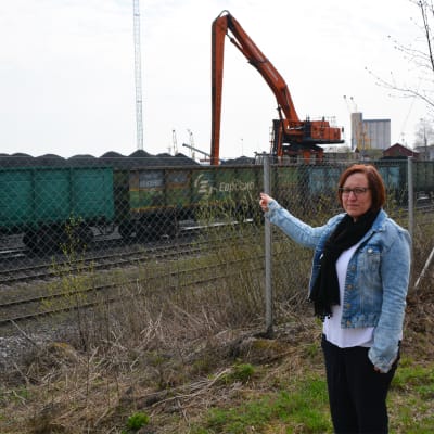 En kvinna står vid ett staket och pekar. Bakom staketet synns tågvagnar, stora kolhögar och en arbetsmaskin.