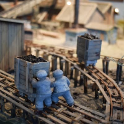 Två små trägubbar skuffar en vagn med kol i en miniatyrmodell av Dalsbruks masugnsområde.