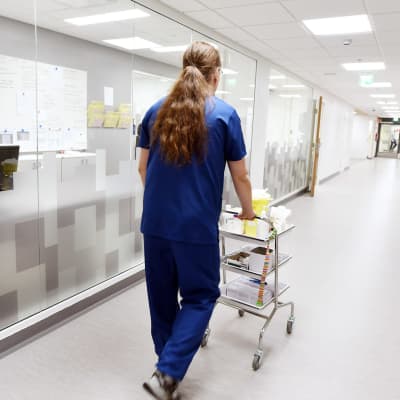 Manlig sjukskötare går i korridor på Mellersta Österbottens Centralsjukhus i Karleby. En kvinnlig anställda sitter bakom en datorskärm i ett intilliggande rum med stora fönster som vetter mot korridoren. 