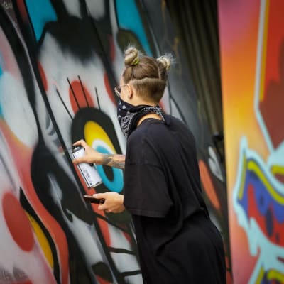 Graffititaiteilija EKA tekee selin kameraan graffititaideteosta värikkäiden seinien keskellä.