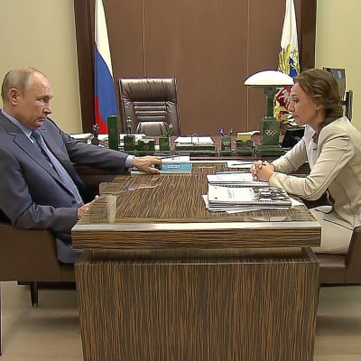 Rysslands president Vladimir Putin och barnombudsman Anna Kuznetsova sitter mittemot varandra vid ett skrivbord och ser allvarliga ut. 