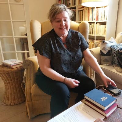 Författaren och historikern Carina Nynäs i sitt hem i Åbo sitter i en gul länstol. Sin vänstra hand har hon på sin mobiltelefon på bordet. Framför henne ligger ett par psalmböcker. 