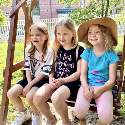 Kolme pikkutyttöä istuvat puisessa puutarhakeinussa kesäisellä kerrostalon pihamaalla.