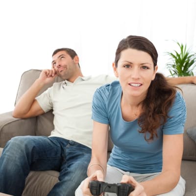 Kvinna och man sitter i soffa, han ser uttråkad ut, hon håller i en spel-kontroller