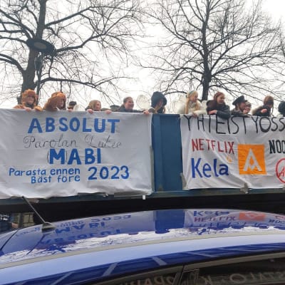 Penkkariasuihin pukeutuneita Parolan lukion abiturientteja seisoo rekan lavalla. Rekan kyljessä on lakanoita, joissa on tekstiä ja kuvia.